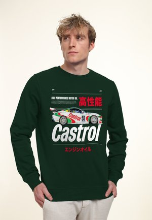 Jeune homme portant un sweat-shirt vert foncé avec un graphisme de voiture de course Castrol et du texte japonais, regardant légèrement vers le haut et sur le côté.