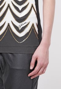 Černé tričko s bílým a zlatým cik-cak vzorem a metalickými akcenty. Spodní lem je zakončený stehem.