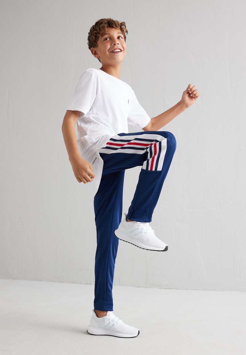 Maglietta bianca a maniche corte, pantaloni blu con strisce rosse e bianche ai lati e sneakers bianche. Bambino in piedi con una gamba alzata.