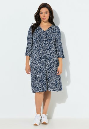 Kvinde iført en navyblå knælængde kjole med hvidt blomsterprint og 3/4-ærmer, kombineret med hvide sneakers, stående mod en ensfarvet baggrund.