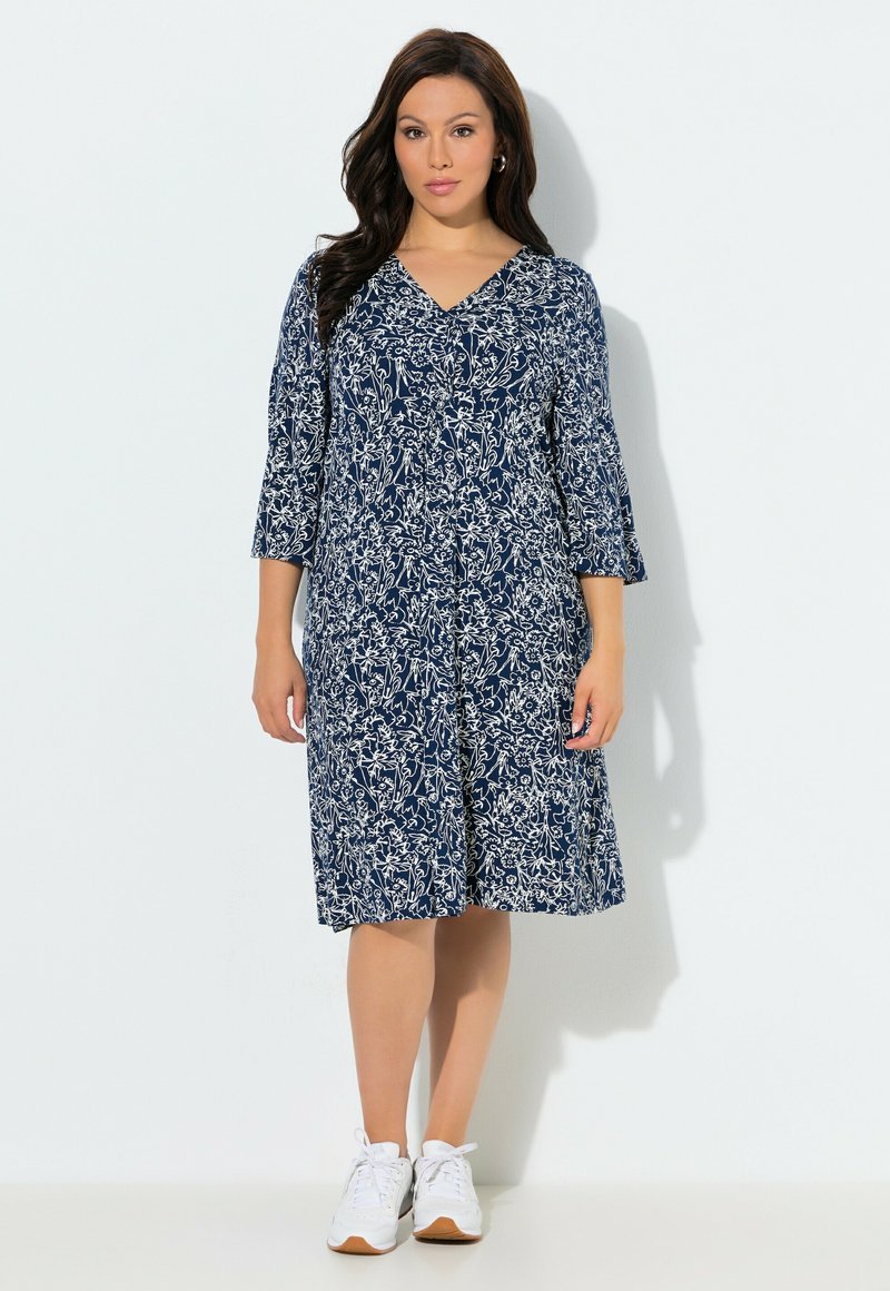 Nainen pukeutuneena laivastonsiniseen, polvipituiseen mekkoon, jossa on valkoinen kukkakuosi ja 3/4-hihat, yhdistettynä valkoisiin tennareihin, seisoo yksiväristä taustaa vasten.