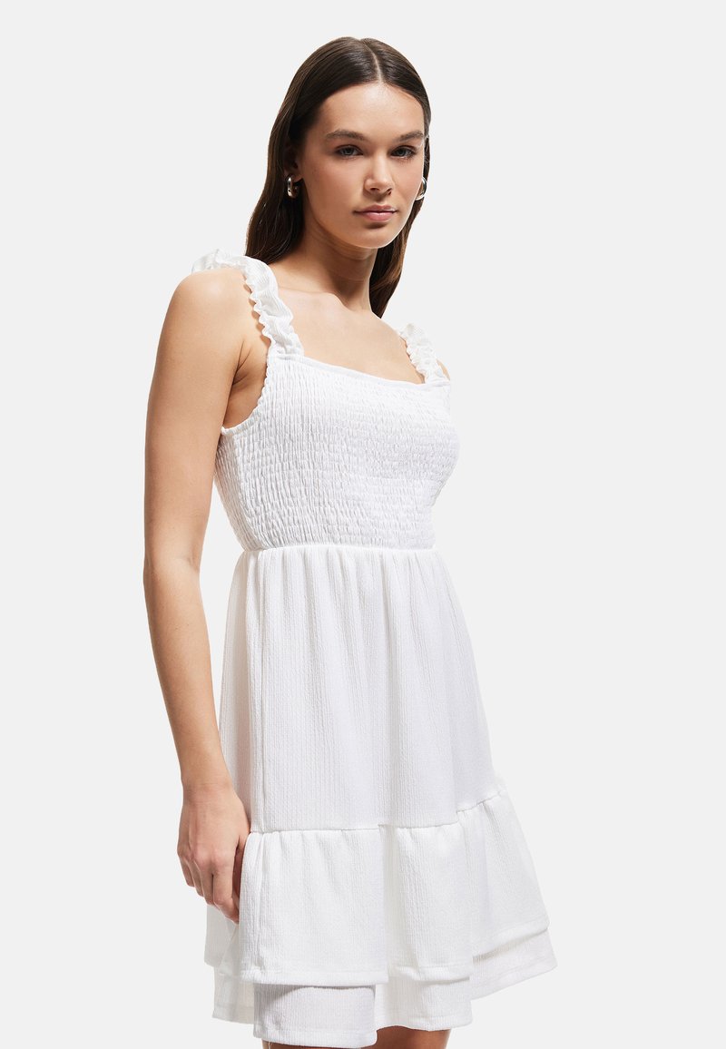 Witte jurk met een vierkante halslijn, gerafelde schouderbanden en elastische smocking bij het lijfje, met een gelaagde rok en textielmateriaal.