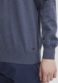 Pulover bleu marin dintr-un material tricotat moale, cu un tiv și manșete ribbate, având un mic eticheta neagră cu logo în partea de jos.