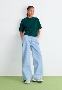 T-shirt oversize vert foncé, jeans larges bleu clair, baskets grises avec des accents verts, et boucles d'oreilles anneaux sur un fond uni.