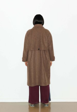 Manteau en laine marron avec une fermeture à boutons dans le dos, manches mi-longues et coupe décontractée, associé à un pantalon large bordeaux et des chaussures à motifs.