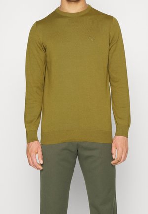 Man draagt een olijfgroene trui met ronde hals en bijpassende groene broek, staand tegen een effen lichte achtergrond.