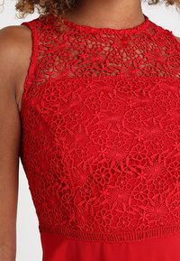 Robe sans manches rouge avec un élégant superposé en dentelle florale sur le buste et un col en dentelle transparent.