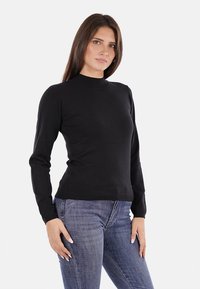 Maglione nero a maniche lunghe con colletto alto e polsini a costine, realizzato in un tessuto liscio. Abbinato a jeans blu aderenti.