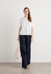 T-shirt blanc à manches courtes, jeans larges foncés, chaussures noires à bout pointu et sac fourre-tout crème, sur fond clair uni.