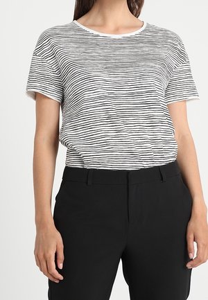 Femme portant une chemise blanche à manches courtes avec de fines rayures horizontales noires, rentrée dans un pantalon noir taille haute.