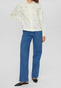 Camicia bianca con ricami floreali colorati, abbinata a jeans blu a gamba dritta e ballerine nere a punta.