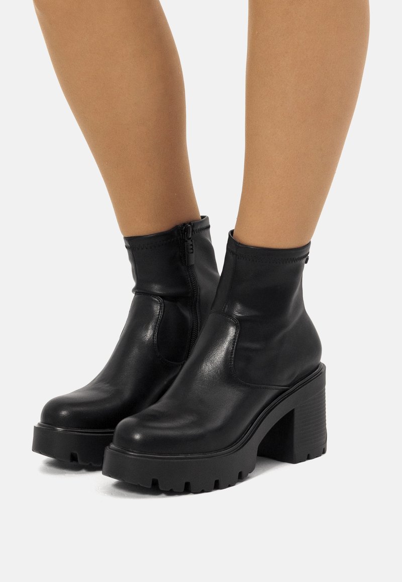 Botas negras de tobillo hechas de cuero suave con una suela de plataforma gruesa y estriada y un zipper lateral. Caño corto con acentos elásticos.