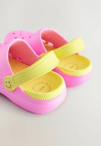 Roze rubberen klompen met gele zolen, voorzien van ventilatiegaten en een brede band met een glimlach-gezicht detail. Getextureerde binnenkant.