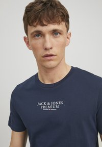 Tamno plava pamučna majica sa okruglim izrezom, s bijelim ispisanim logotipom "JACK & JONES PREMIUM" na prsima. Kratki rukavi.