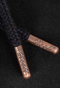 Primer plano de cordones negros trenzados con terminales metálicas de bronce grabadas con la palabra "Freedom" sobre un fondo de tela oscura.
