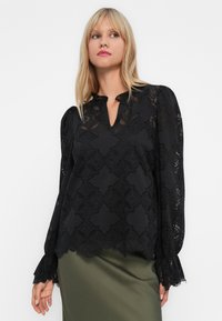 Femme blonde portant un haut en dentelle noire à manches longues avec des bords festonnés et une jupe en satin verte, regardant légèrement sur le côté.