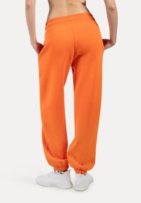 Orange sweatpants med en afslappet pasform, ribbet talje, og elastiske manchetter, parret med hvide sneakers. Blødt materiale, minimalistisk design.