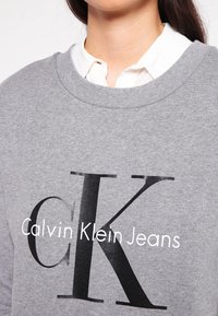 Sweatshirt grigio con scollatura tonda, caratterizzato da un grande "CK" nero e "Calvin Klein Jeans" in testo bianco sul petto. Tessuto morbido.