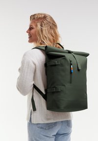 Mochila verde de tipo roll-top con correas ajustables, bolsillo con cremallera y cierres a presión. Textura suave, diseño minimalista y detalle del logo en la parte inferior.