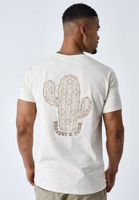 Crème-kleurig T-shirt met een geborduurd cactussenontwerp op de achterkant in bruin en witte patronen. Korte mouwen, ronde halslijn.
