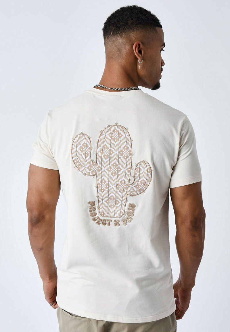 Camiseta color crema con un diseño de cactus bordado en la parte posterior en patrones marrones y blancos. Mangas cortas, cuello redondo.