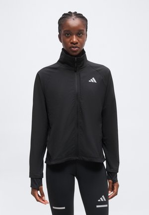 Jeune femme portant une veste de sport noire zippée et des leggings noirs avec un logo blanc et des bandes réfléchissantes sur fond uni.