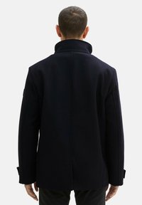 Granatowy wełniany płaszcz z wysokim kołnierzem, prostym krojem i bocznymi kieszeniami; posiada przeszywane szwy i zapinane mankiety. Pokazano widok z tyłu.