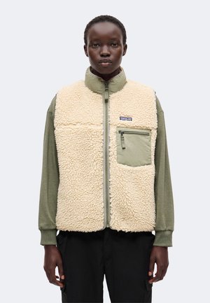 Person trägt eine cremefarbene Fleeceweste mit Reißverschlusstasche auf der Brust über einem grünen Langarmshirt und schwarzen Hosen vor weißem Hintergrund.