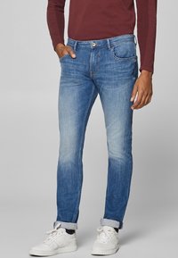 Sinivihreät farkut slim fit -mallissa, joissa on haalistunut pinta, viisi taskua ja nappikiinnitys, yhdistettynä valkoisiin tennareihin.