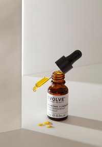 Bärnstensfärgad glasdroppflaska med etikett "Evolve Organic Beauty" och svart lock, innehållande gul olja och intilliggande gula droppar.