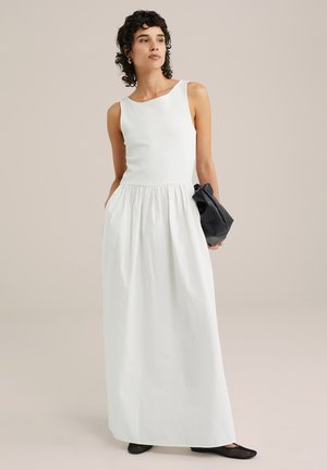 Witte mouwloze jurk met een ribbenstructuur bovenkant en volumineuze rok, met een gathered taille. Model houdt een zwarte handtas vast en draagt zwarte schoenen.