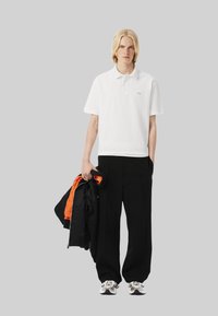 Vit pikétröja med korta ärmar, kompletterad med lösa svarta byxor. Modellen håller en svart jacka med orange foder och bär mönstrade sneakers.