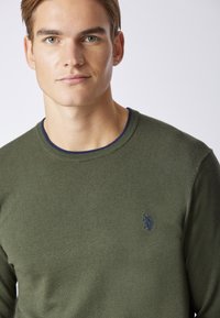 Maglione verde lavorato a maglia con colletto rotondo e bordi blu navy. Presenta un piccolo logo ricamato sul petto. Texture liscia, design aderente.