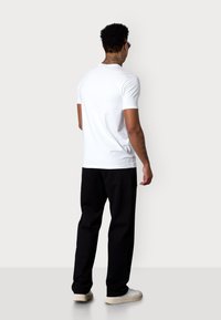 T-shirt blanche à manches courtes, en coton, au design ajusté. Associée à un pantalon noir et des baskets de couleur claire, vue de dos.