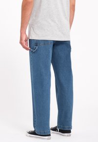 Jeans in denim blu con una vestibilità rilassata, dotati di tasca posteriore e dettagli laterali, abbinati a sneakers bianche e nere.