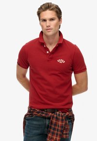 Polo rouge en coton avec col, deux boutons et un logo blanc "SUPER DRY". Associé à une chemise en flanelle à carreaux nouée à la taille.