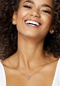 Collier en or rose avec un pendentif en forme de cœur ornée de trois petites pierres précieuses claires. Le modèle a les cheveux bouclés et porte des boucles d'oreilles assorties.