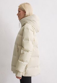 Beige pufferjack met capuchon, met een gestikte structuur en zijzakken met rits. Zachte, gladde stof met een volumineuze silhouet.