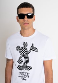 Camiseta blanca de algodón con un gráfico de Mickey Mouse en blanco y negro con un diseño a cuadros. Se llevan gafas de sol, lo que resalta el look casual del conjunto.