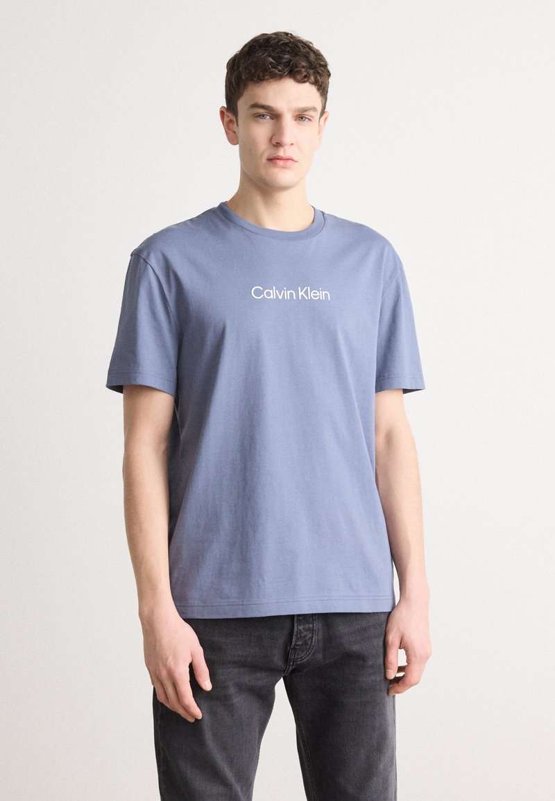 Zils kokvilnas T-krekls ar apaļu kakla izgriezumu un īsām piedurknēm. Pazīmes baltais "Calvin Klein" logos uz krūšu. Saskaņots ar tumši pelēkiem džinsiem.
