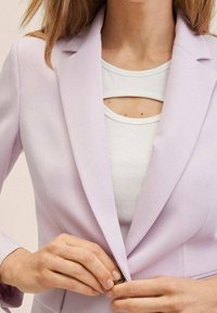 Jasnofioletowy żakiet o dopasowanym kroju, z klapami i pojedynczym zapięciem na guzik, nałożony na biały, prążkowany top z wycięciem.