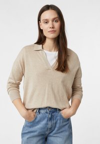 Beżowy sweter z dzianiny z dekoltem w serek i kołnierzykiem; długie rękawy i ściągacze na mankietach. Noszony na białej koszulce z luźnymi niebieskimi dżinsami.