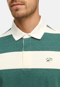 Camisa polo a rayas verdes y crema con cuello y cierre de botón. Presenta un logotipo bordado con el detalle "EST. 1993".