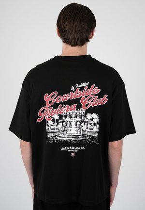 Mand iført en sort T-shirt med rød og hvid tekst "Courtside Riviera Club" og en illustration af en herregård med palmer og en fontæne.