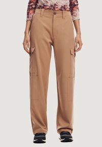 Pantaloni cargo beige con vestibilità rilassata, dotati di quattro tasche, chiusura con bottone e una superficie in tessuto liscia. Indossati con sneaker nere.