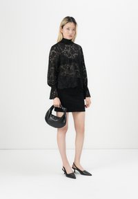 Svart blondebluse med høy hals og voluminøse ermer, kombinert med et svart miniskjørt, svarte slingback-sko og en liten svart håndveske.