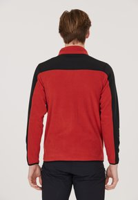 Röd fleecjacka med svarta accenter; har en krage, långa ärmar och en mjuk textur. Bakifrån framhäver kontrasterande färger.