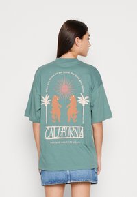 Teal oversized t-shirt, short sleeves, features a graphic of a sun, leopards, and palm trees, with "CALIFORNIA" in bold lettering below.