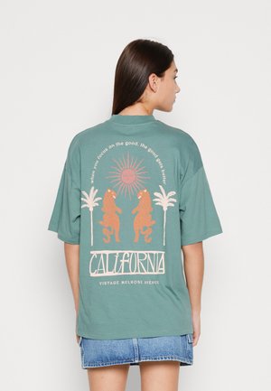 Türkisfarbenes Oversized-T-Shirt mit kurzen Ärmeln, das ein Grafikdesign mit einer Sonne, Leoparden und Palmen zeigt, darunter in fetter Schrift "CALIFORNIA".