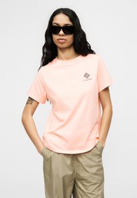 Γυναίκα που φοράει μαύρα γυαλιά ηλίου, ροδακινί μπλούζα Columbia και μπεζ παντελόνι με τα χέρια στις τσέπες, στέκεται μπροστά σε λευκό φόντο.